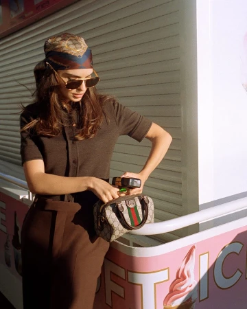 Model wearing a monogram headscarf and sunglasses holding a Gucci monogram handbag against a shuttered kiosk