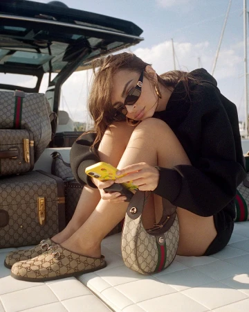 Woman in dark sunglasses holding a phone while seated on a boat surrounded by monogram luggage for a Gucci campaign.