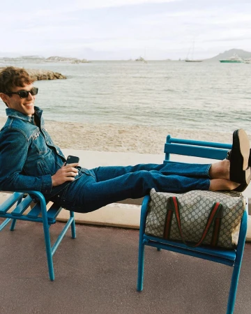 Kit Butler wearing denim jacket and jeans resting feet on a blue chair with a Gucci monogram duffel bag by the sea