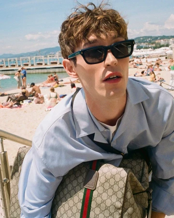 Man wearing sunglasses and light blue shirt carrying a monogram canvas bag on a sunny beach