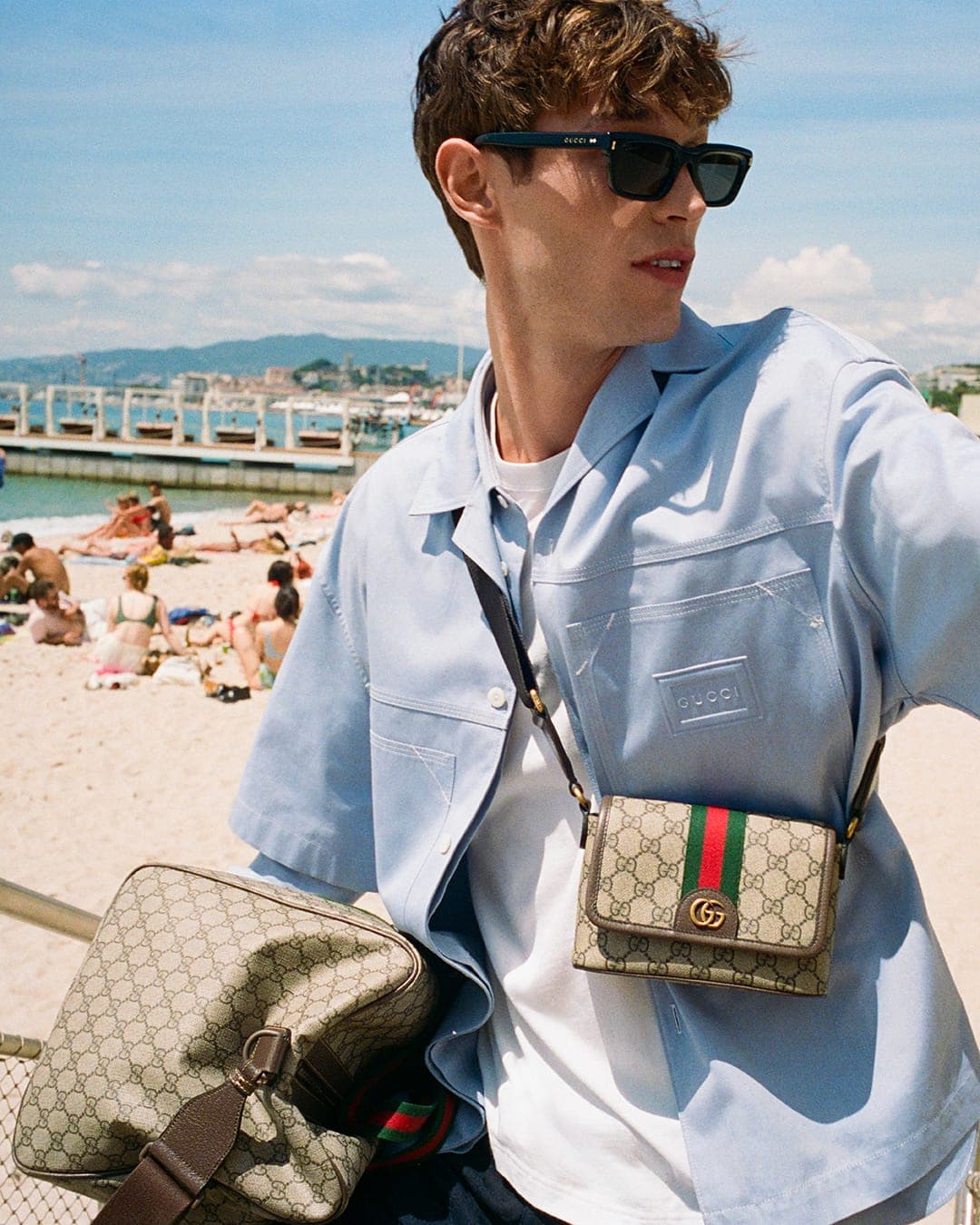 Kit Butler wearing sunglasses and a light blue shirt carrying GG monogram bags on a sunny beach.