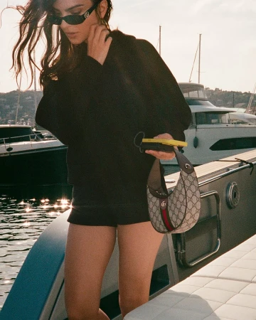 Emily Ratajkowski wearing sunglasses and holding a Gucci monogram handbag on a boat deck.