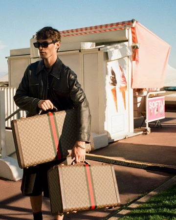 Man wearing sunglasses and a black leather jacket walking past an ice cream stand while carrying monogram luggage cases