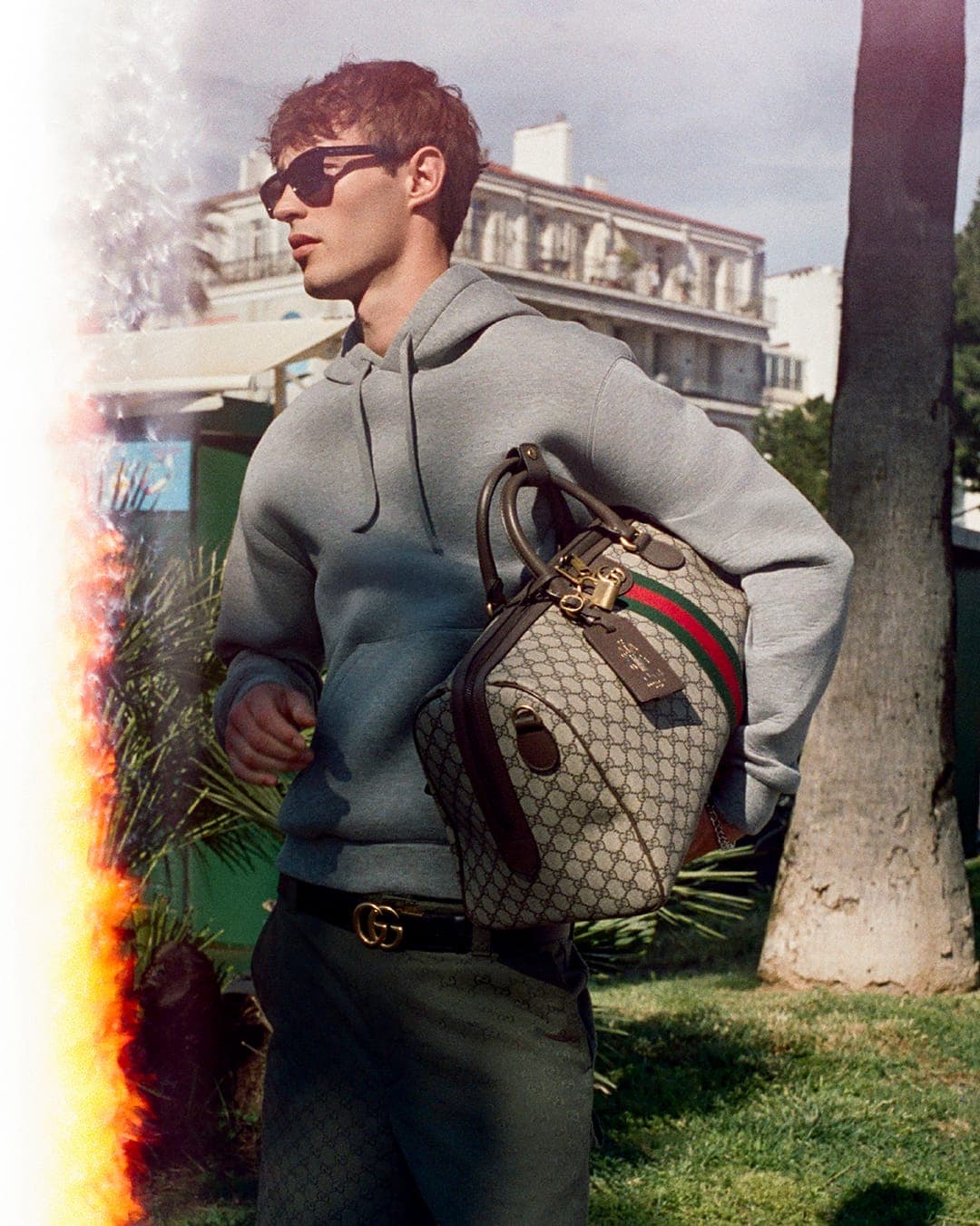 Kit Butler wearing a grey hoodie and sunglasses holding a monogram handbag outdoors for Gucci GG Monogram