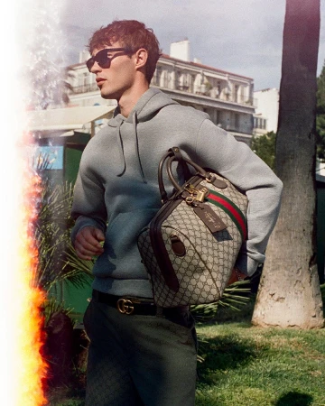 Kit Butler wearing a grey hoodie and sunglasses holding a monogram handbag outdoors for Gucci GG Monogram