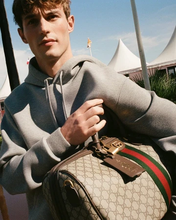 Kit Butler wearing a grey hoodie holding a Gucci monogram duffle bag outdoors under a clear blue sky.