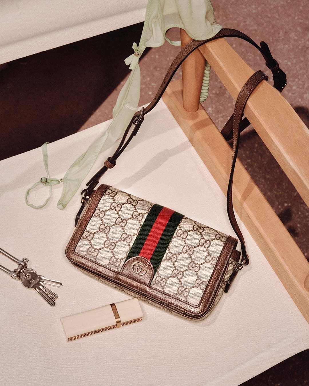 Gucci monogram shoulder bag resting on a white surface alongside keys, lipstick, and a green scarf draped over a wooden chair.
