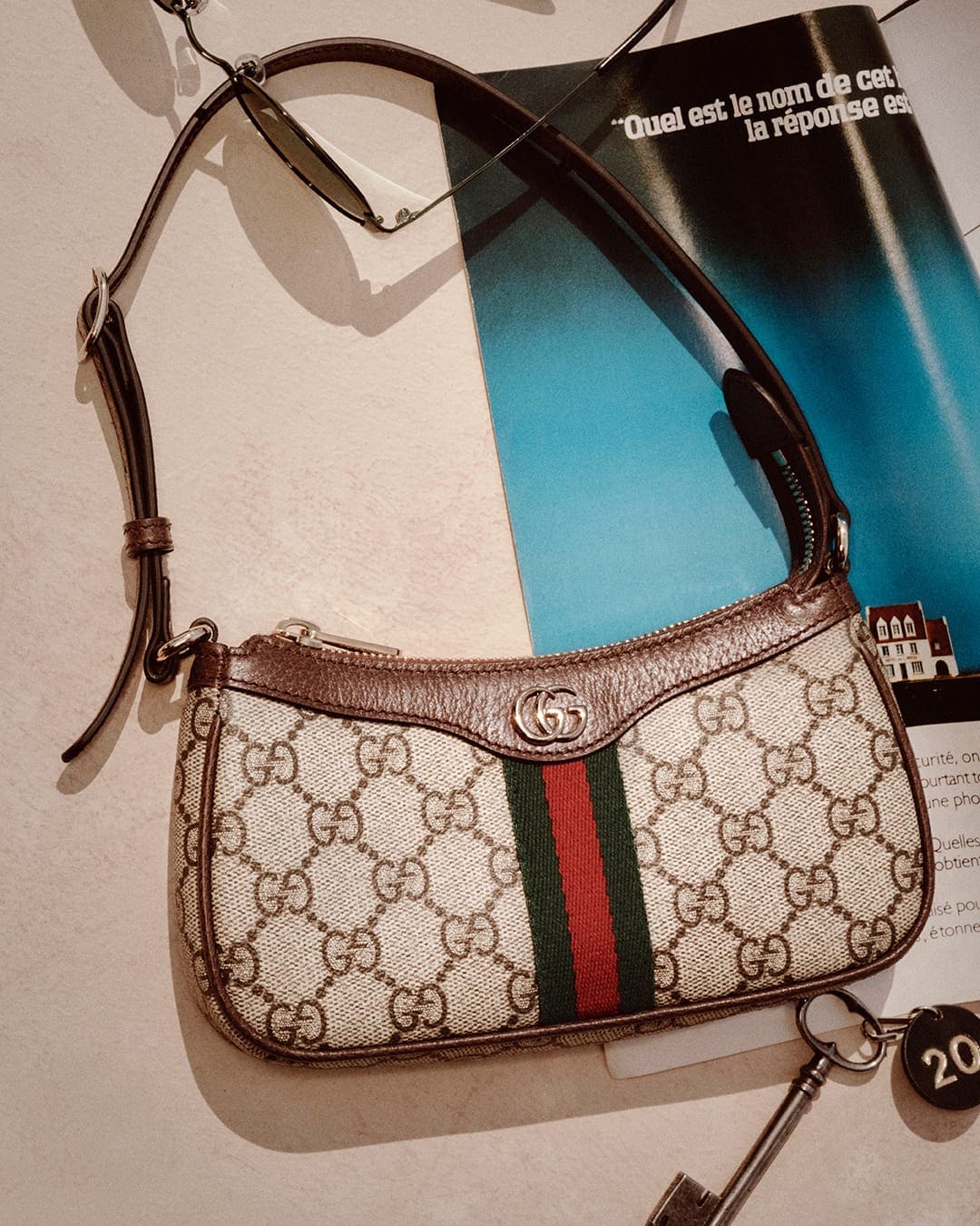Gucci monogram shoulder bag with green and red stripe resting on a flat surface next to sunglasses and a key