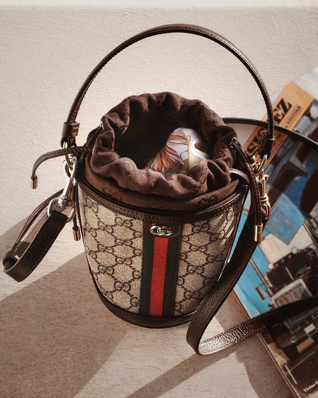 Gucci monogram bucket bag with brown leather trim and red-and-green web stripe resting on a neutral surface