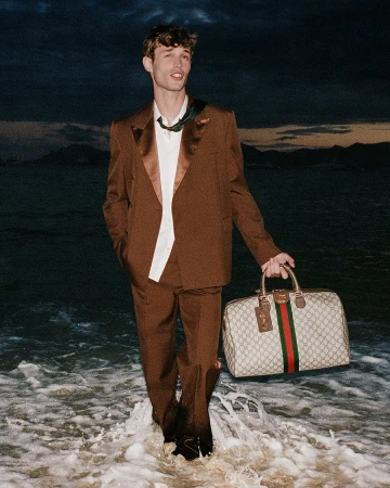 Man wearing a brown suit holding a monogram duffel bag while standing in ocean surf at dusk