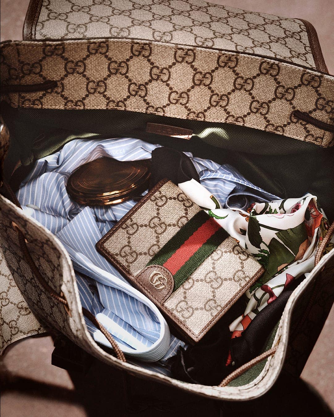 Open monogram canvas backpack packed with a striped shirt, patterned silk scarf, and small wallet with red and green stripes.