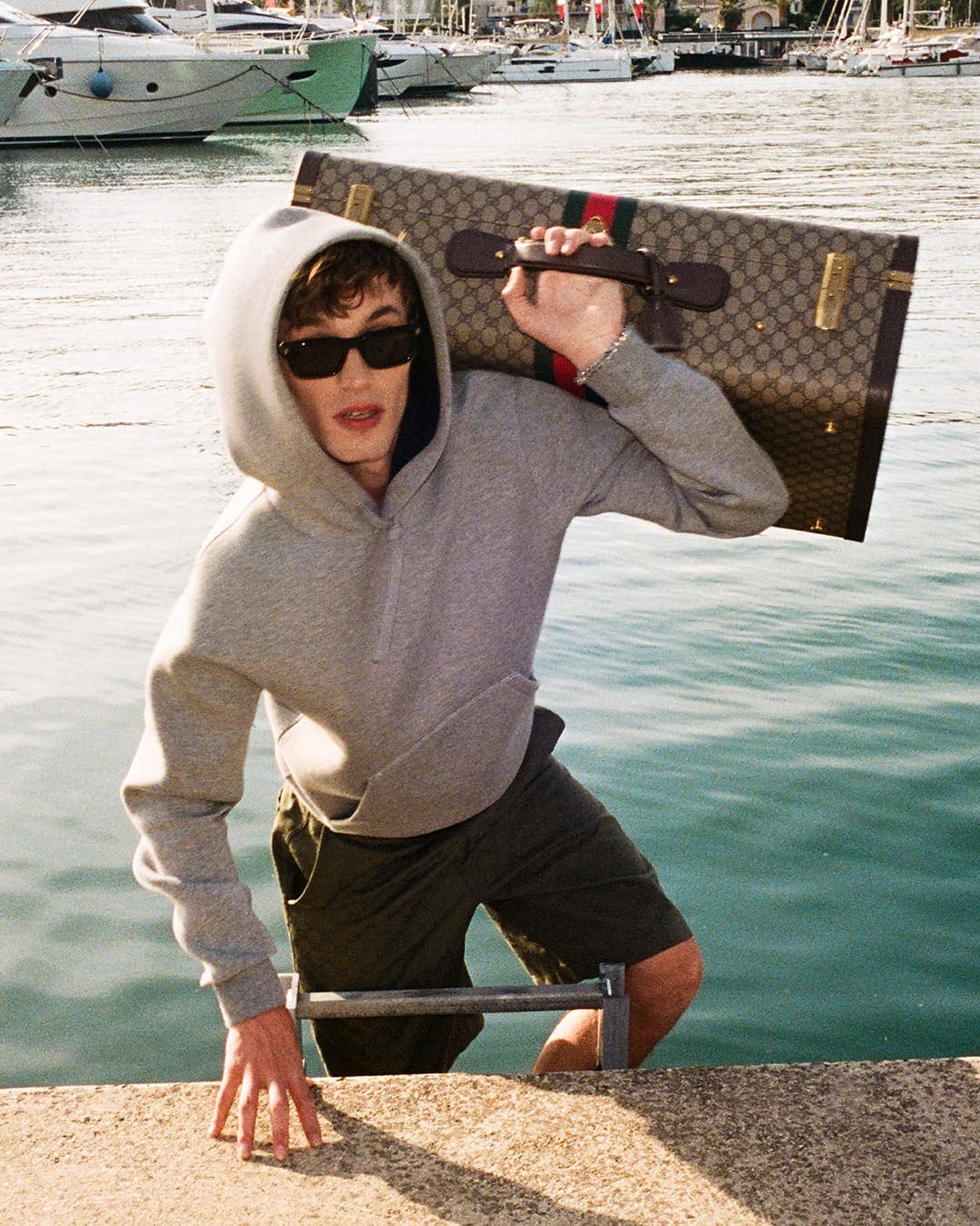Man wearing a grey hoodie and sunglasses carrying a monogram suitcase over his shoulder near a marina.