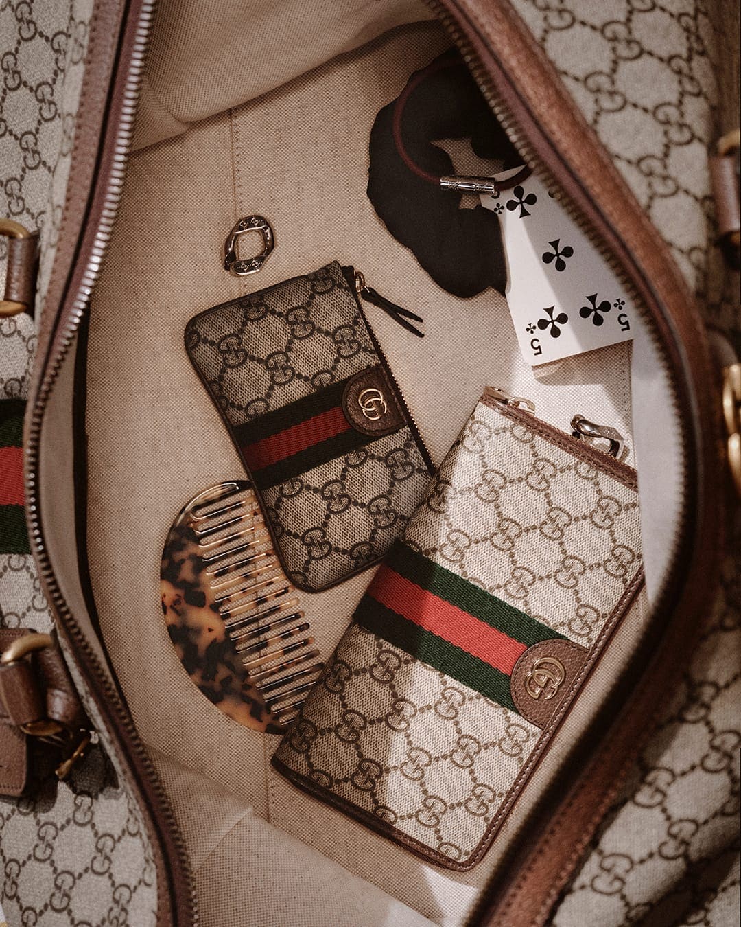 Overhead interior shot of a large Gucci monogram canvas bag filled with accessories including wallets, a tortoiseshell comb, and playing cards.