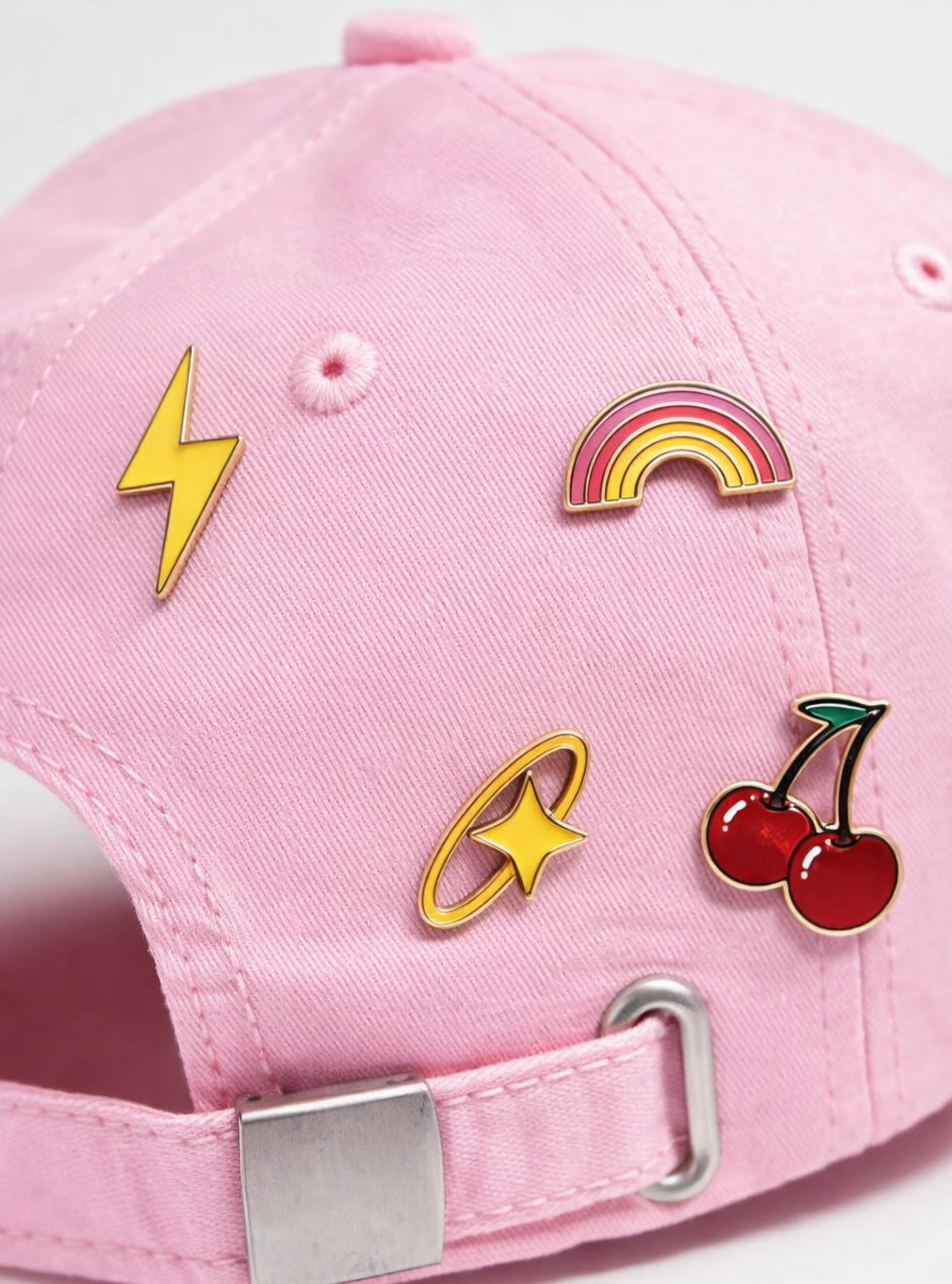 Close-up of a pink cap adorned with colorful enamel pins including a lightning bolt, rainbow, star, and cherries.