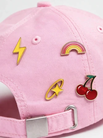 Close-up of a pink cap adorned with colorful enamel pins including a lightning bolt, rainbow, star, and cherries.