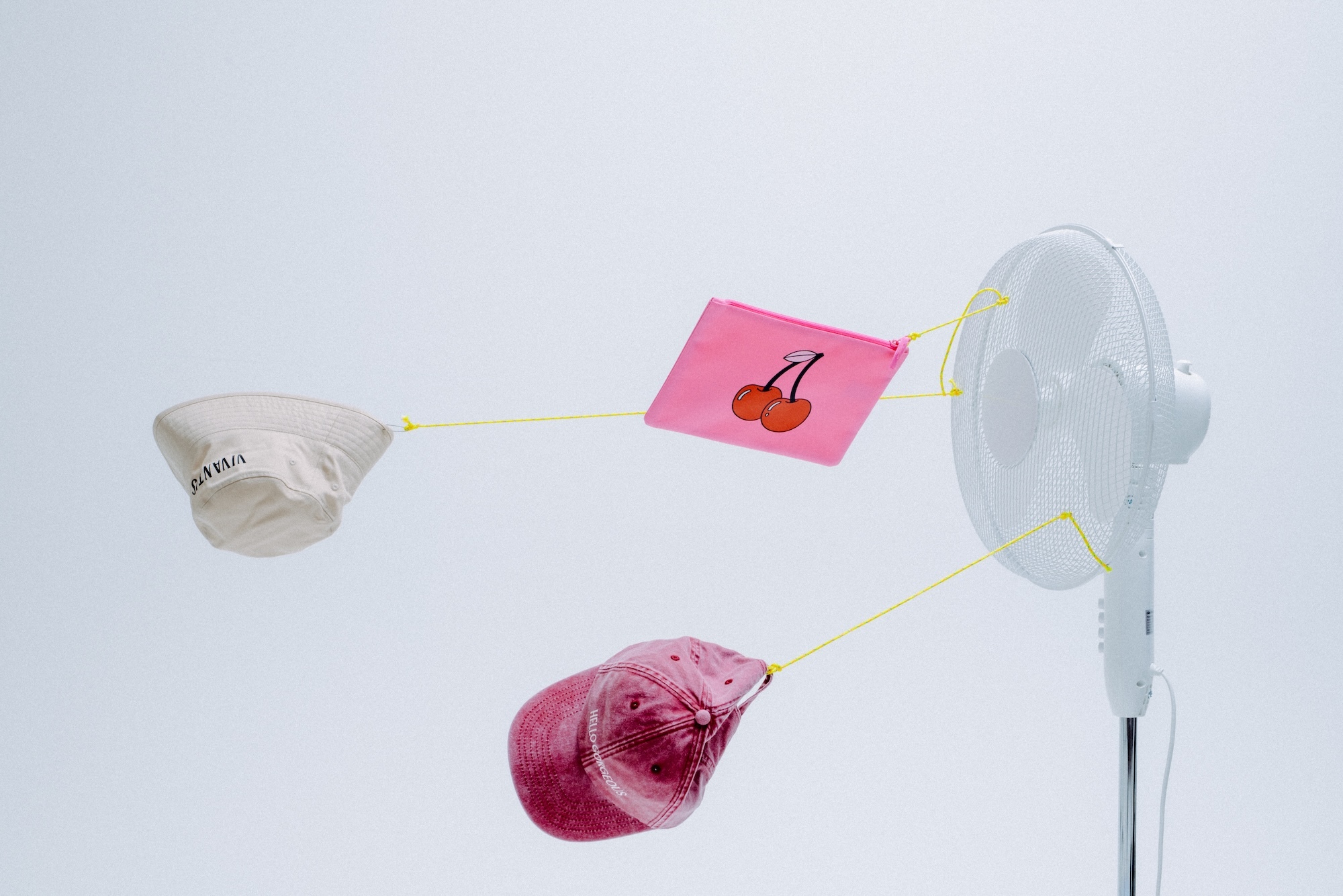 White electric fan blowing accessories and hats attached by yellow strings against a clean background