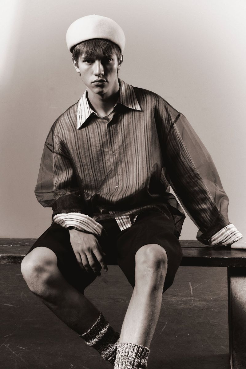 Monochrome portrait of a young adult man in an open collared shirt, shorts, cap, and socks, sitting on a ledge in studio lighting.
