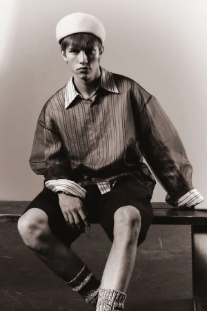 Monochrome portrait of a young adult man in an open collared shirt, shorts, cap, and socks, sitting on a ledge in studio lighting.