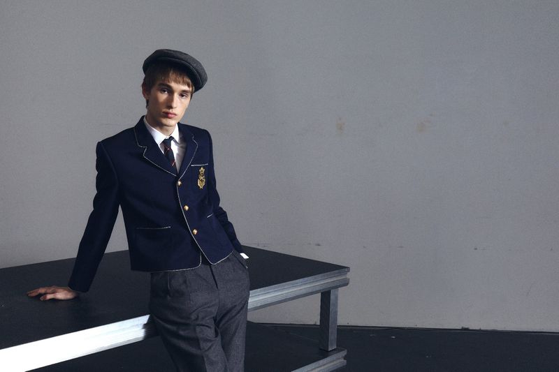Young man in school uniform with newsboy cap posing in a studio, leaning on a black table against a grey backdrop. Fashion editorial style.