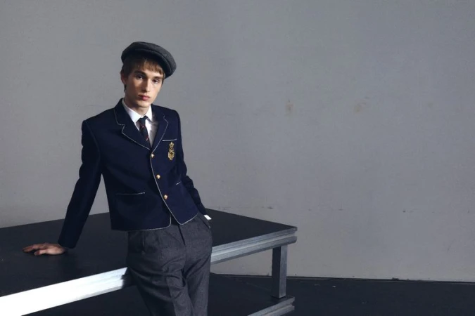 Young man in school uniform with newsboy cap posing in a studio, leaning on a black table against a grey backdrop. Fashion editorial style.