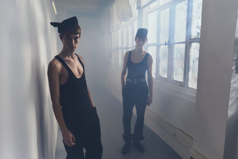 Two young adult men in black tank tops and hats pose in a hallway with natural light from nearby windows, creating a hazy, diffused atmosphere.