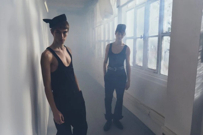 Two young adult men in black tank tops and hats pose in a hallway with natural light from nearby windows, creating a hazy, diffused atmosphere.