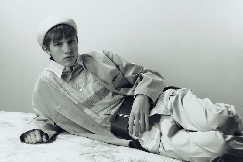 Black and white studio portrait of a young adult male model with dark hair wearing a light-colored jacket, striped shirt, pants, and beret.