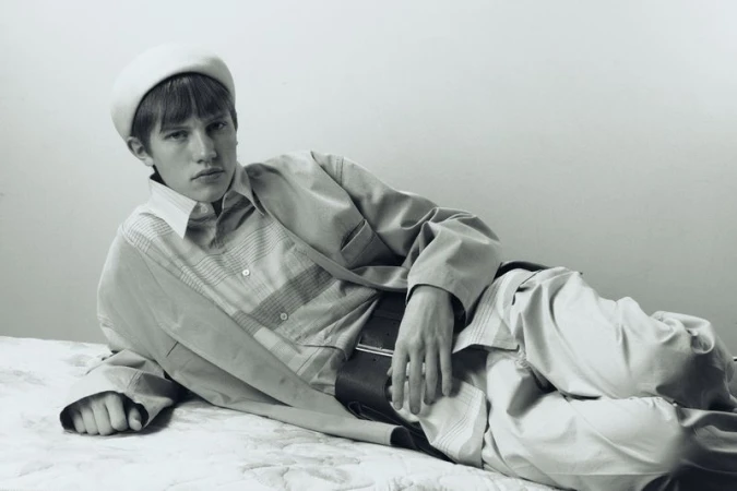 Black and white studio portrait of a young adult male model with dark hair wearing a light-colored jacket, striped shirt, pants, and beret.