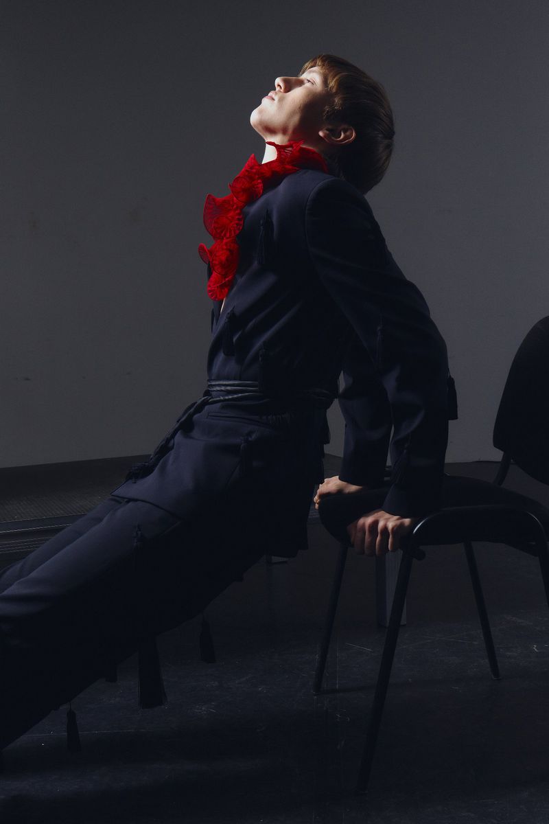 Adult woman in a black suit poses in a studio environment while leaning on a chair with a red feathered boa around her neck.
