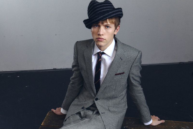 Young man in a grey suit and a striped hat sits on a wooden surface against a light grey wall in a studio setting, looking at the camera.