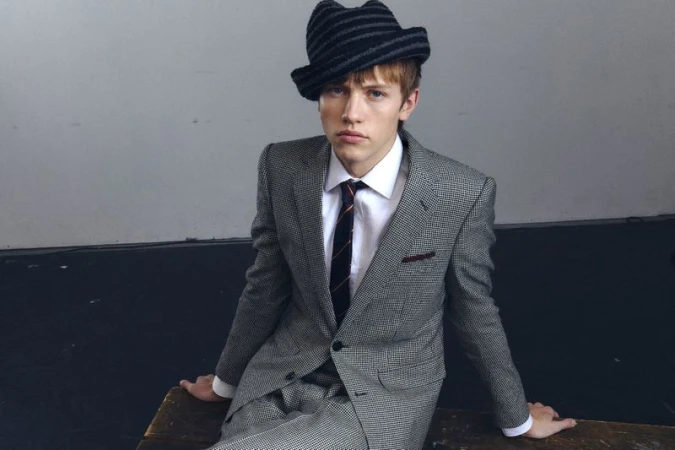 Young man in a grey suit and a striped hat sits on a wooden surface against a light grey wall in a studio setting, looking at the camera.