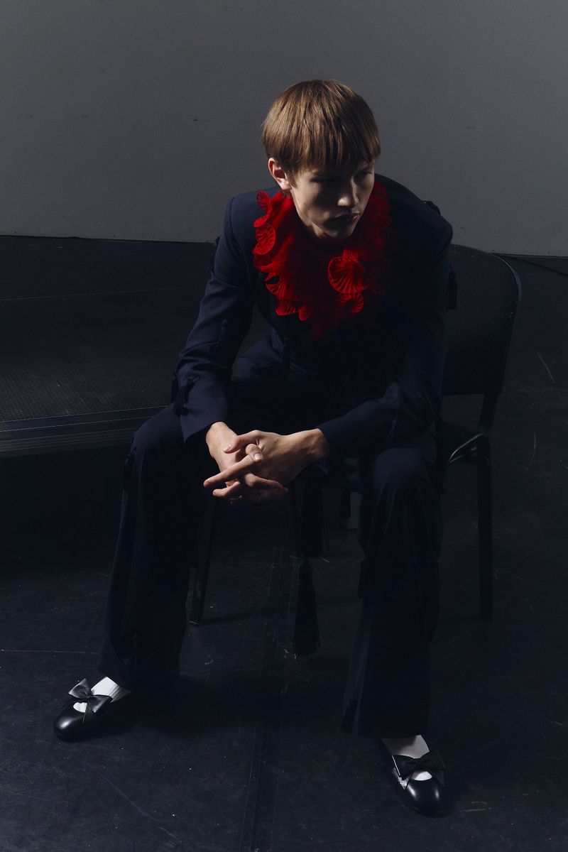 A young adult seated on a black chair, wearing a dark suit and red ruffled collar, contrasted against a dark background with moody lighting.
