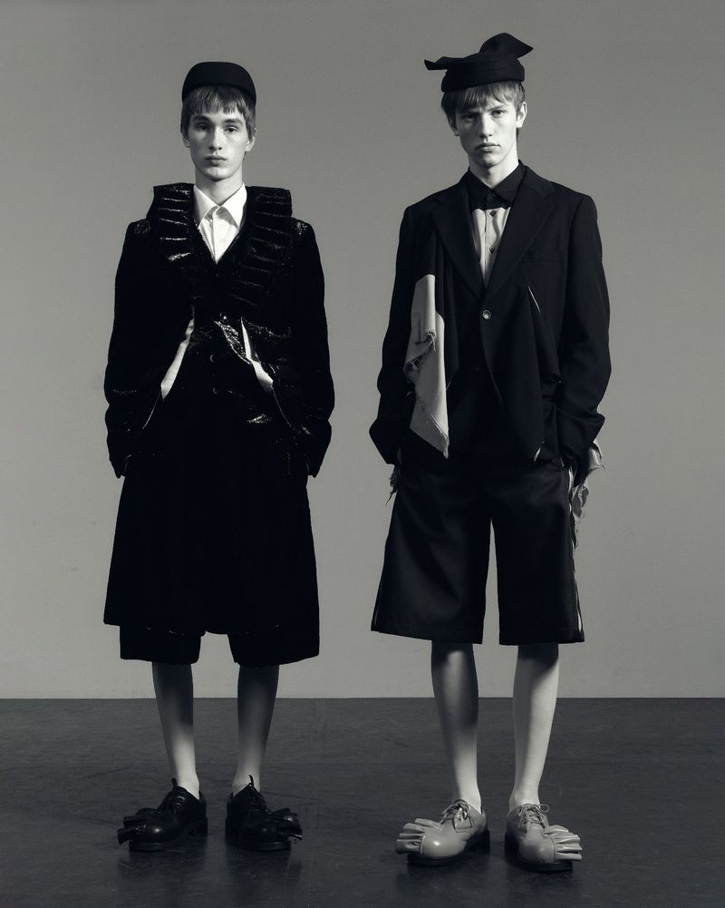 Two young male models in black and white studio shot. One wears a structured jacket and shorts, the other a suit with a drape.