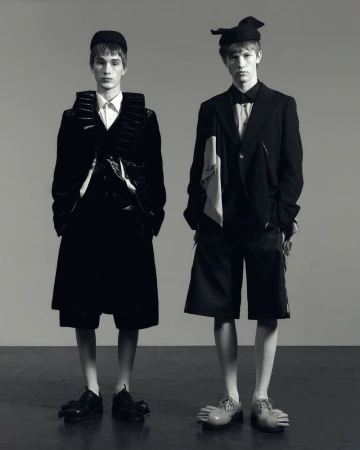 Two young male models in black and white studio shot. One wears a structured jacket and shorts, the other a suit with a drape.