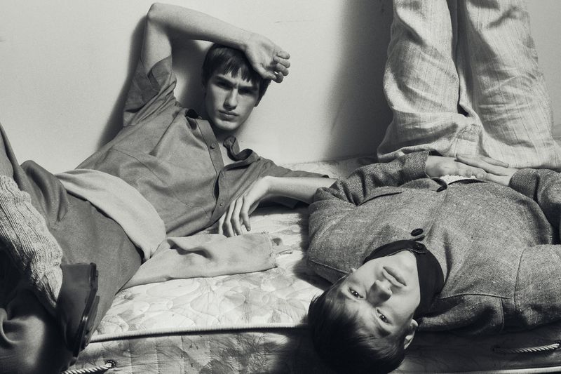 Two men in vintage style clothes lie side by side on a mattress in a dimly lit, black and white photograph.