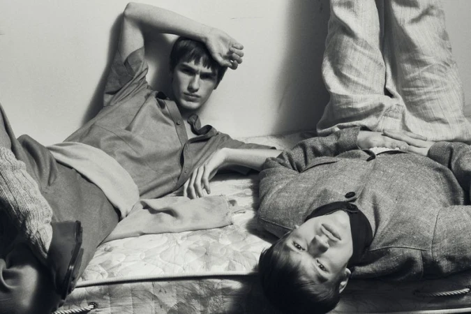 Two men in vintage style clothes lie side by side on a mattress in a dimly lit, black and white photograph.