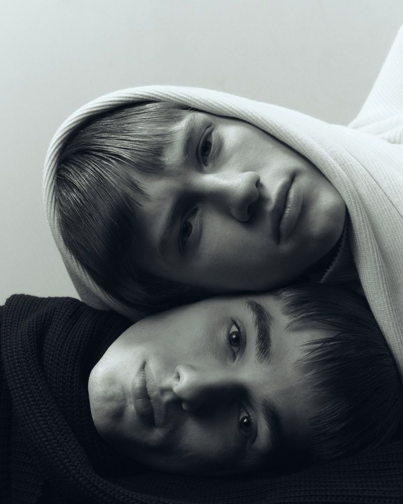 Monochromatic portrait of two young people, one wearing a hooded garment, close together in a fine art portrait style.
