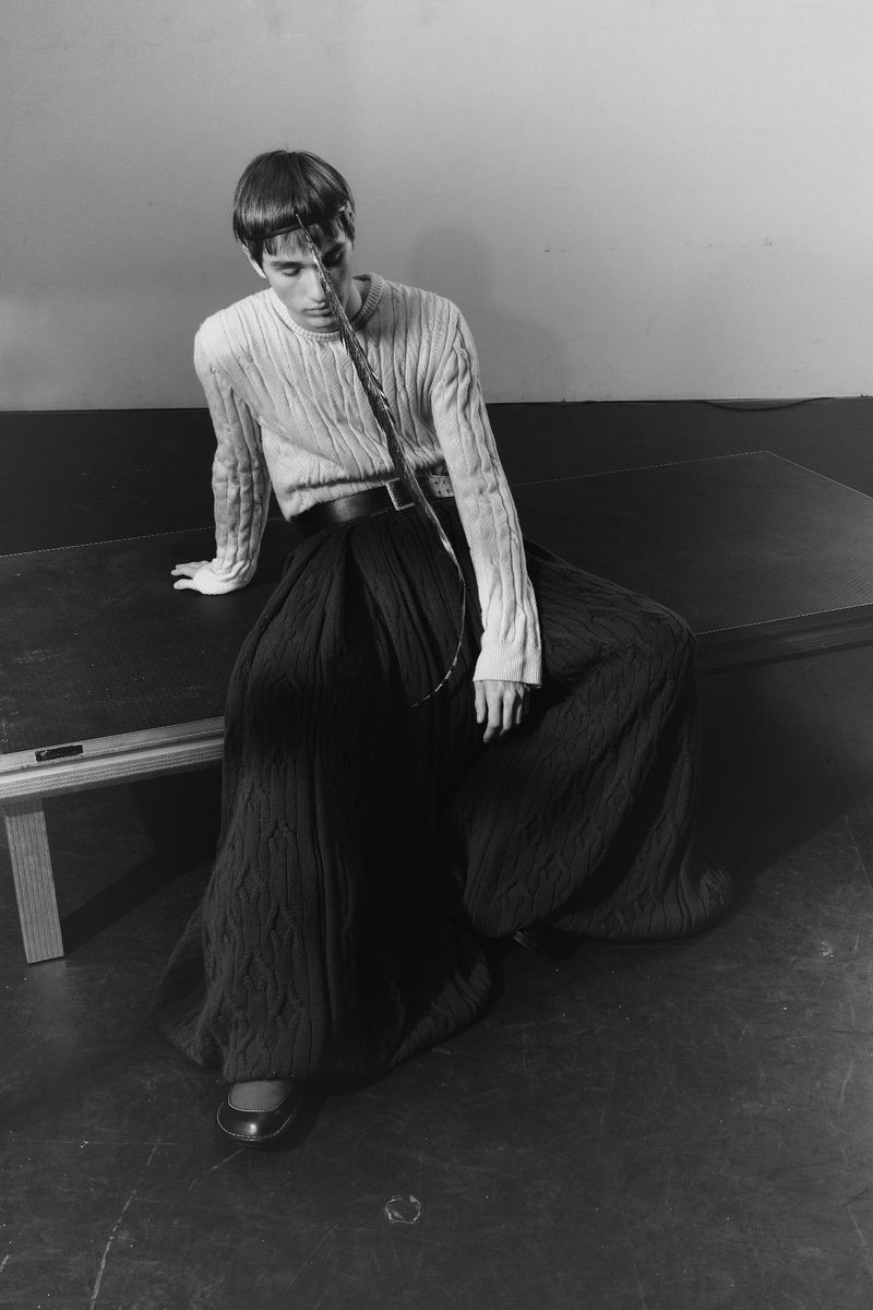 Monochromatic fashion shot of a slender person with short hair seated on a table, wearing a long-sleeved top and wide pleated skirt.