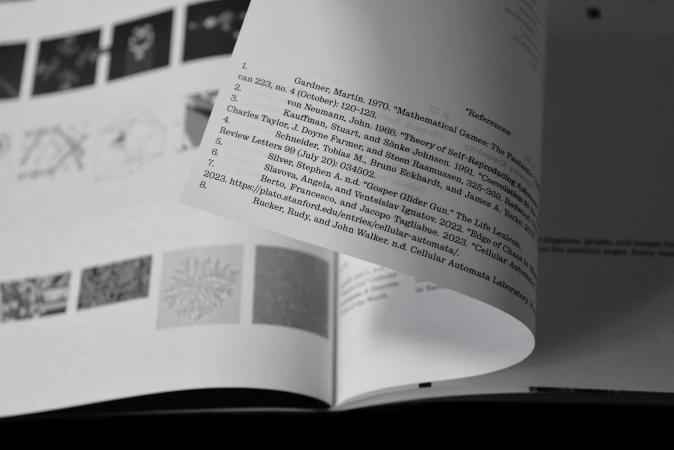 Close-up monochrome view of a curved book page with printed text and reference lists, set against a background of small diagrams.