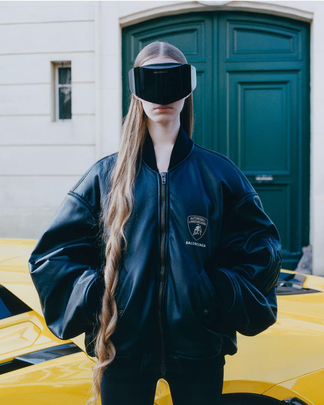 Model wearing a black leather bomber jacket and futuristic visor standing in front of a green door and yellow car