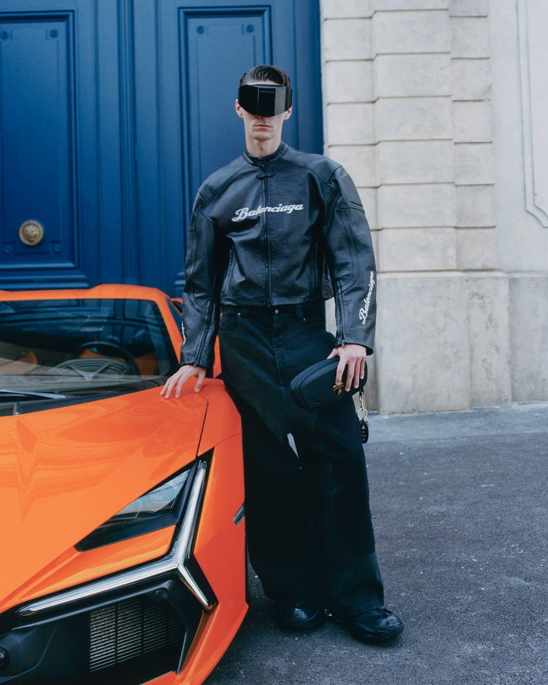 Model wearing black leather jacket and visor sunglasses standing next to an orange sports car before a blue door