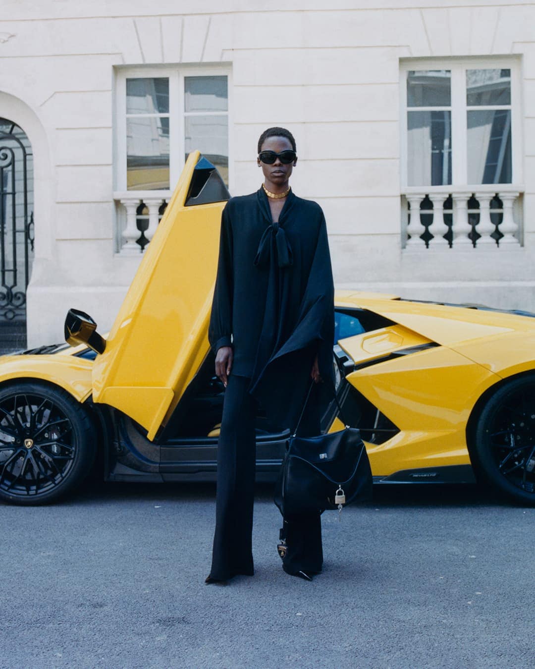 Model in black clothing and sunglasses standing before a yellow sports car with an open door