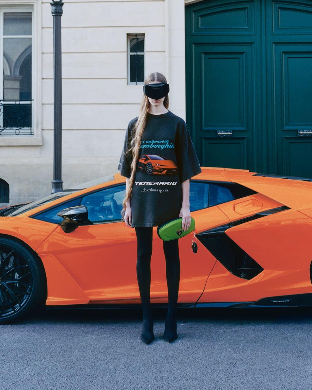 Model wearing dark visor and graphic t-shirt standing before an orange sports car and large teal doors.