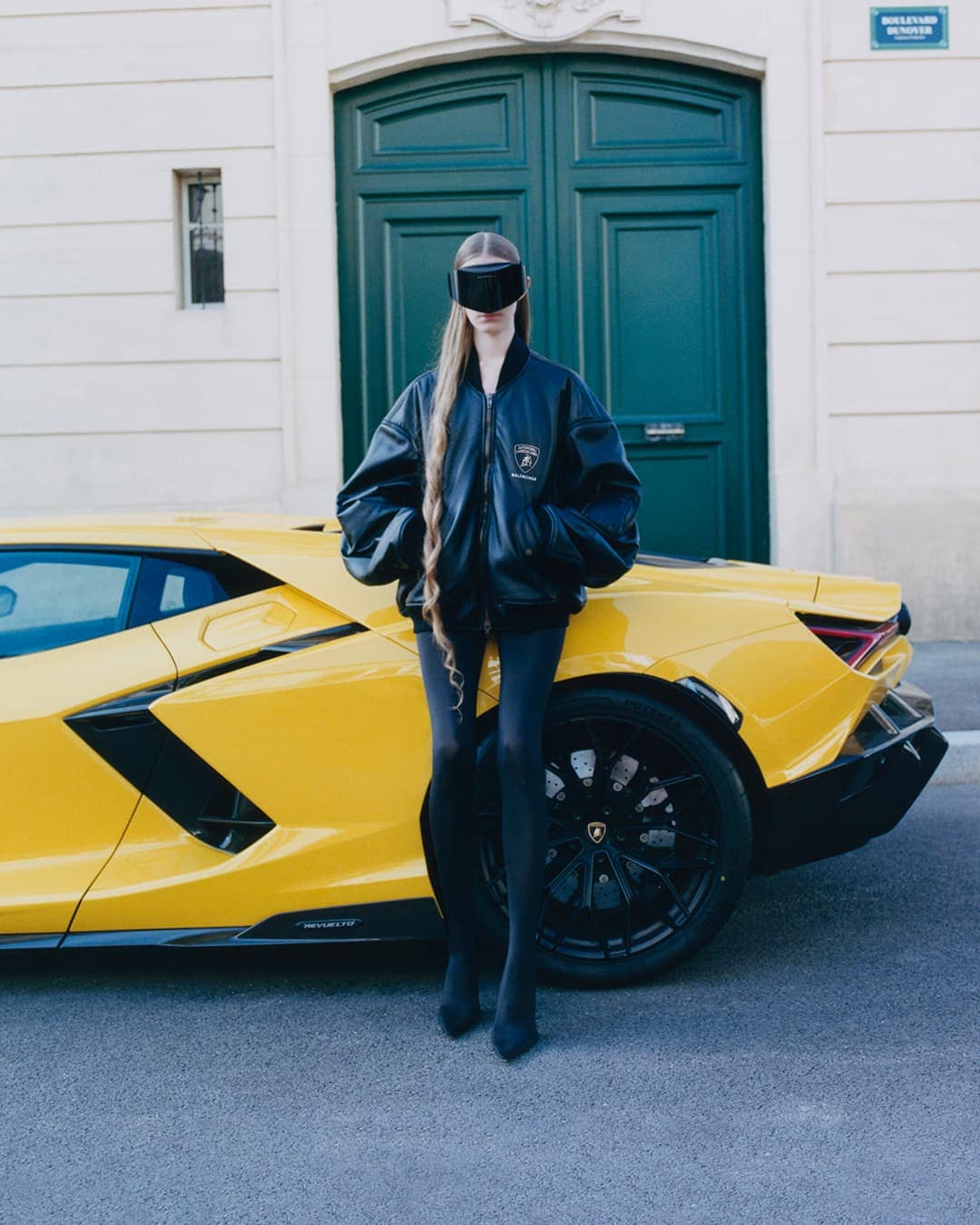 Model wearing dark visor and black leather jacket leaning against a yellow sports car on a street