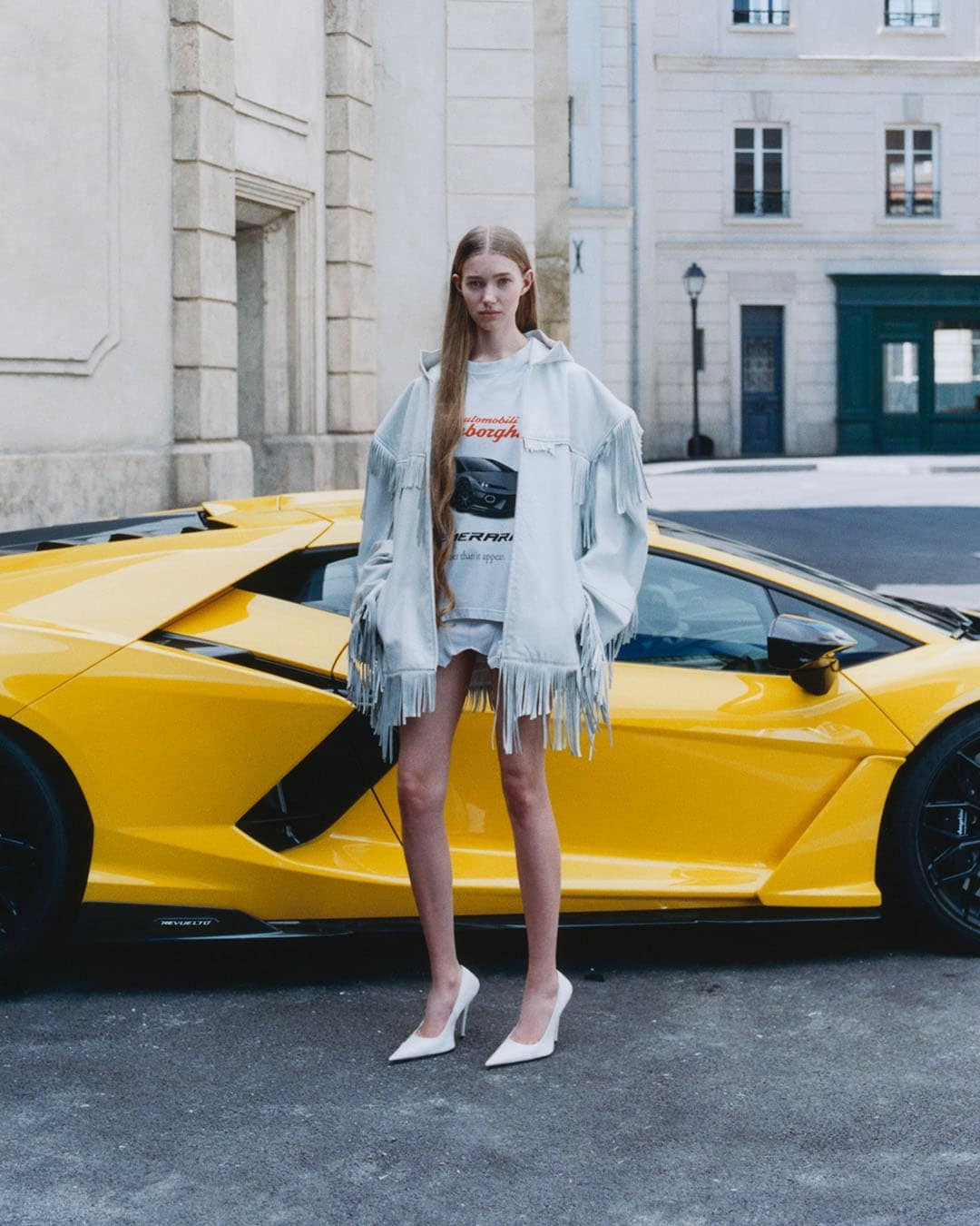 Model with long blonde hair wearing a fringed jacket standing in front of a bright yellow sports car on a city street
