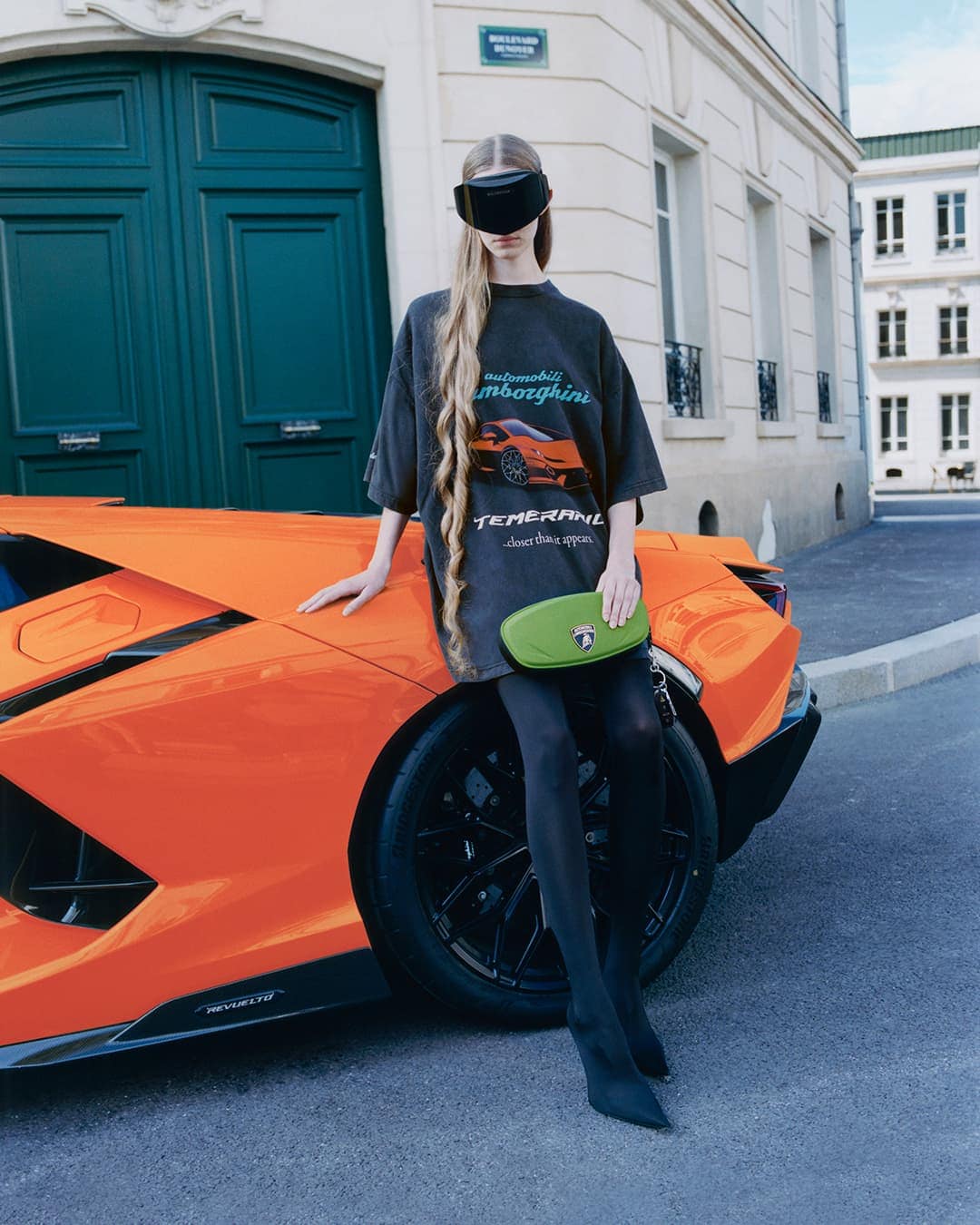 Model wearing dark eyewear and oversized graphic t-shirt leaning against a vibrant orange sports car on a city street.