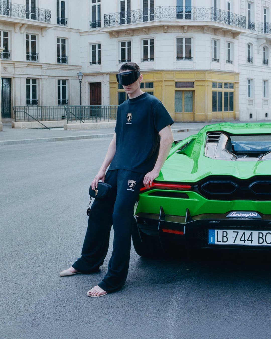 Man wearing dark visor and matching sweatsuit leaning against a vibrant green sports car on a European city street.