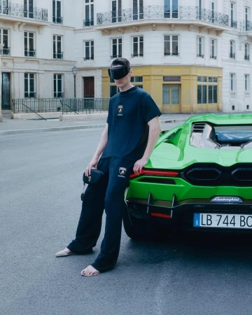 Man wearing dark visor and matching sweatsuit leaning against a vibrant green sports car on a European city street.