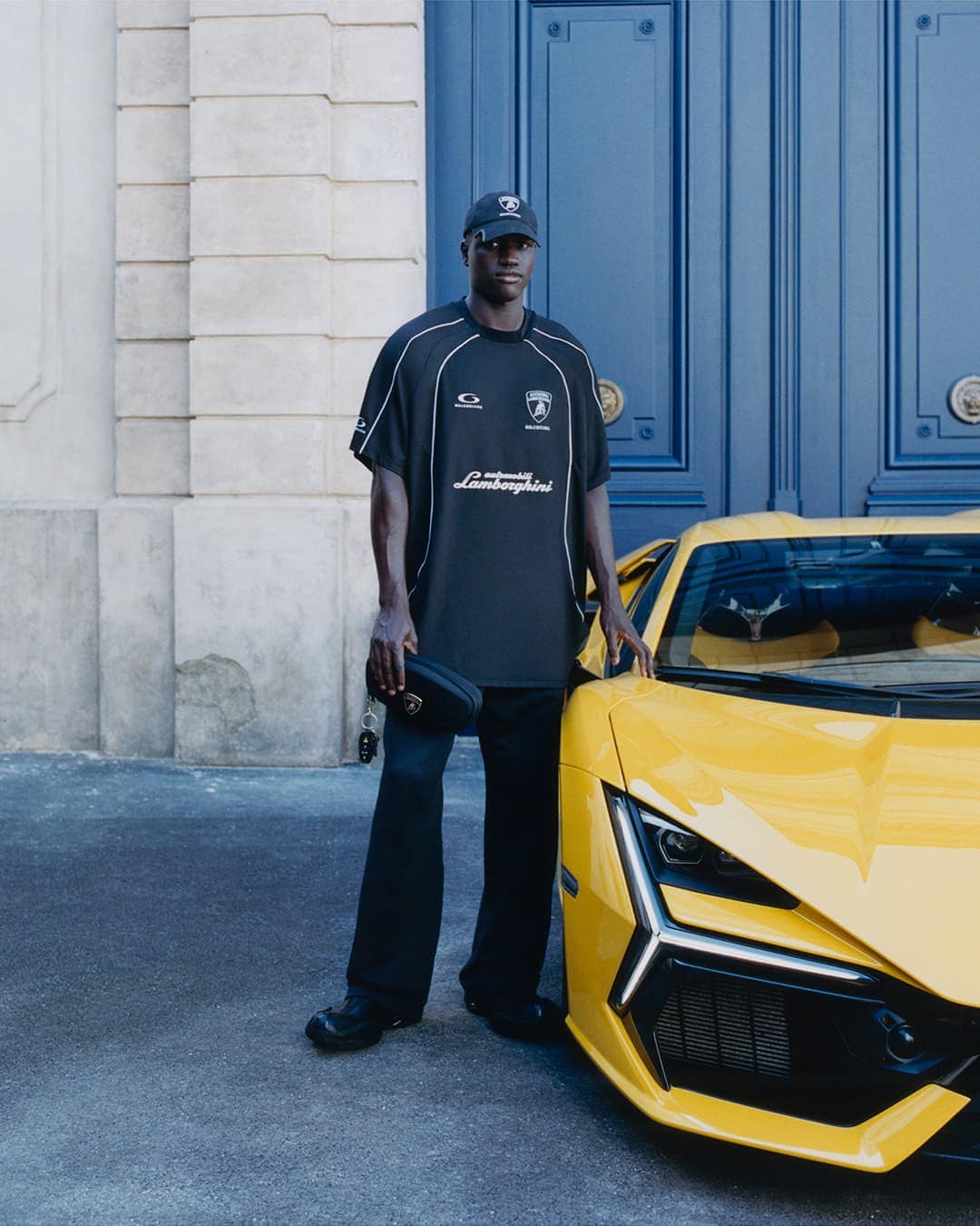 Tall man in black cap and oversized t-shirt standing beside a bright yellow sports car in front of a blue doorway