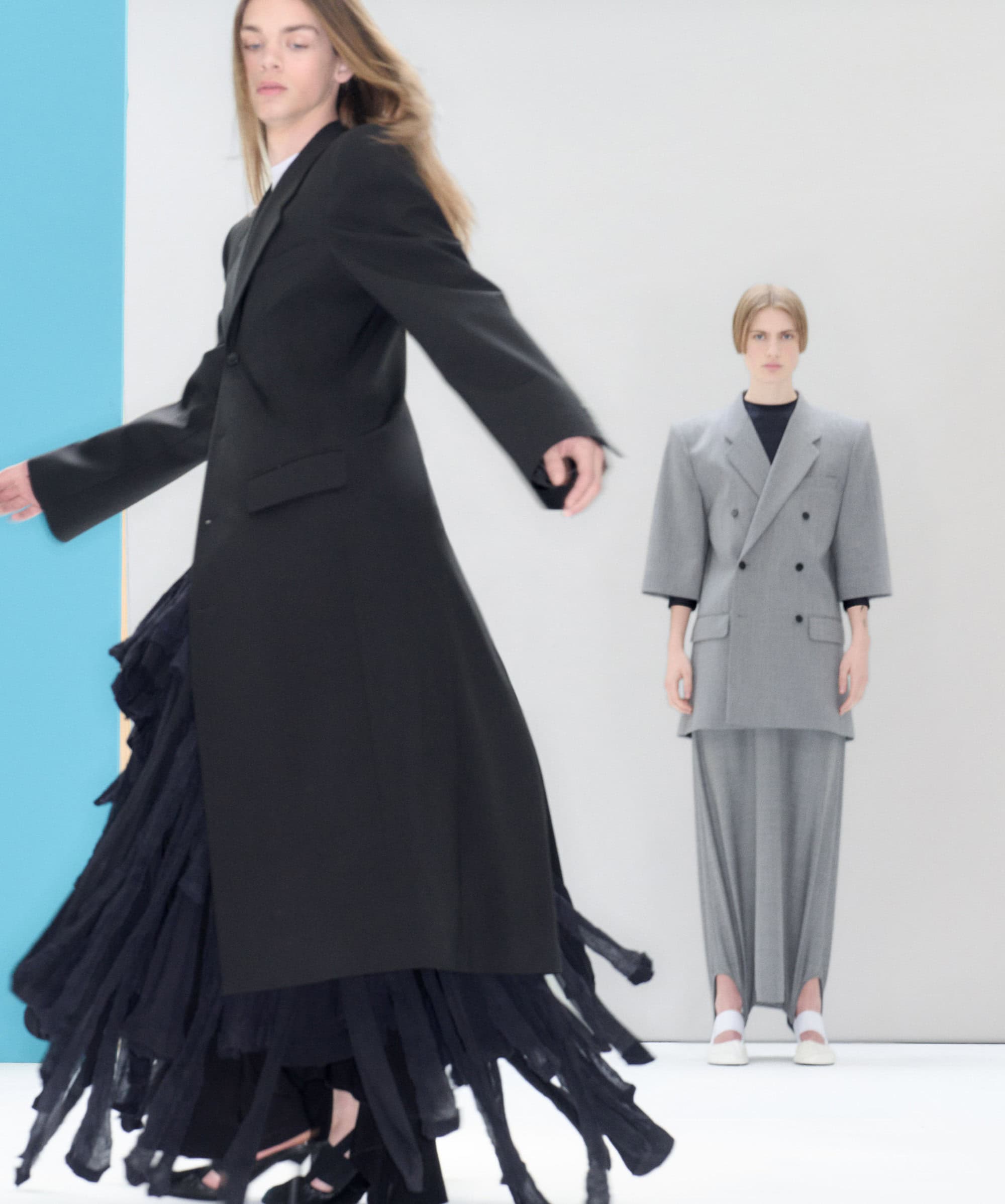 Two models showcasing Alainpaul Spring 2025 collection against a minimalist studio background with blue and white vertical panels.
